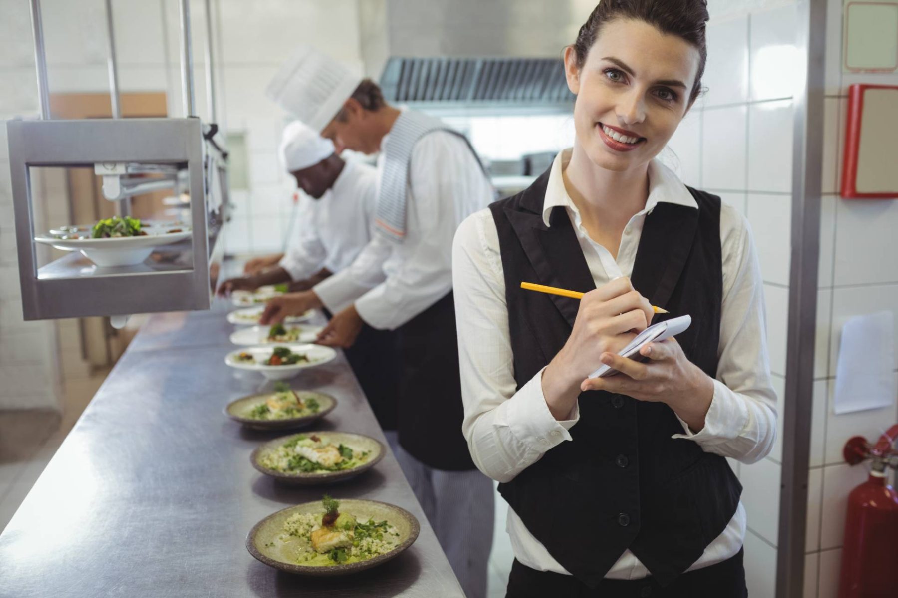 Smiling server holding a notepad and pencil in a restaurant kitchen, with chefs plating dishes in the background.