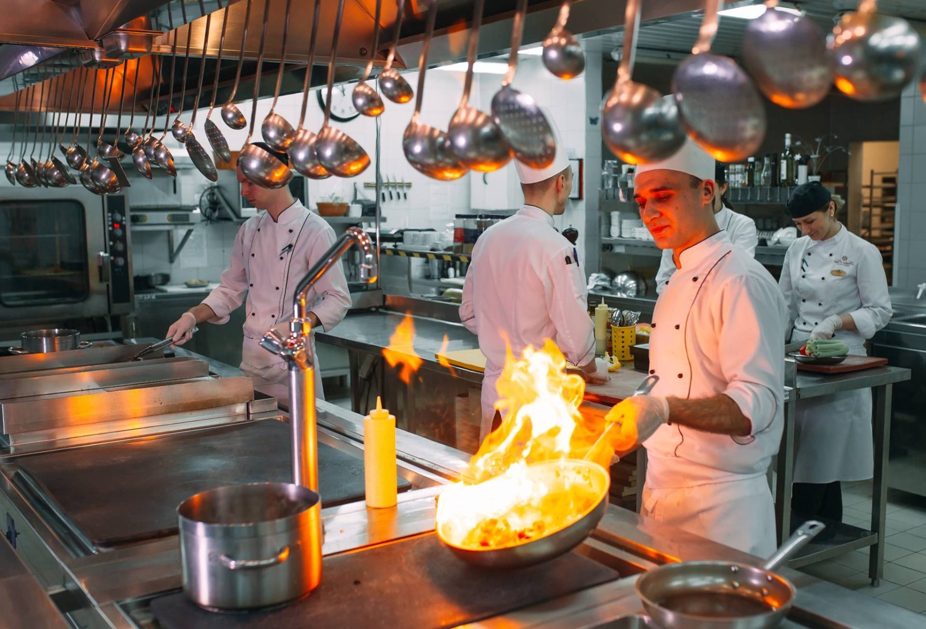 Chefs working in a commercial kitchen; front cook safely flambés a pan under hanging ladles.