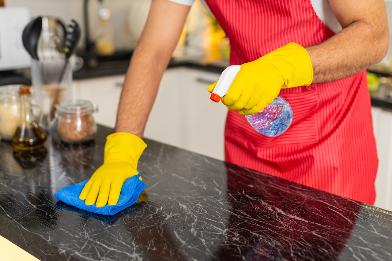 Person in a home kitchen wearing gloves disinfecting a countertop with spray cleaner and a cloth.