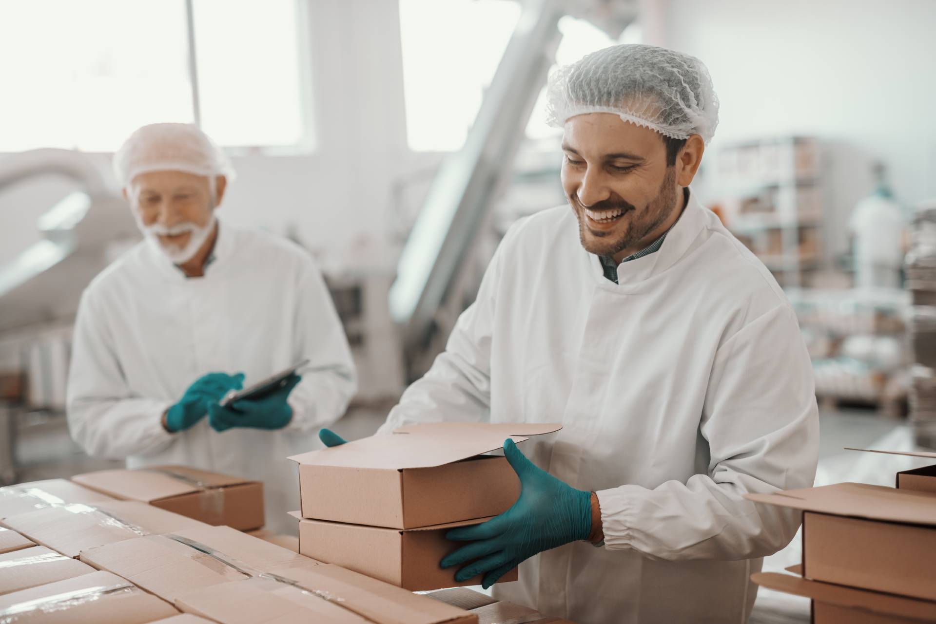 Food production workers in hairnets and gloves packing and stacking cardboard food boxes on a processing line.