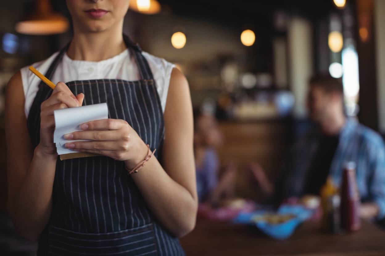 Restaurant server in an apron writing an order on a notepad in a busy dining room.