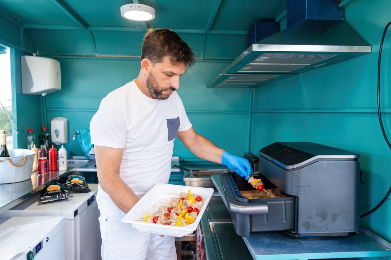 Chef wearing gloves, placing skewers of meat and vegetables into a countertop air fryer inside a small commercial kitchen.