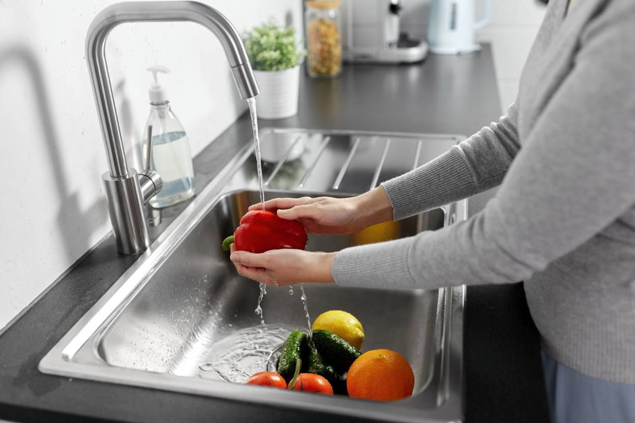 Home cook washing fresh vegetables under running water in a kitchen sink as part of safe food handling practices