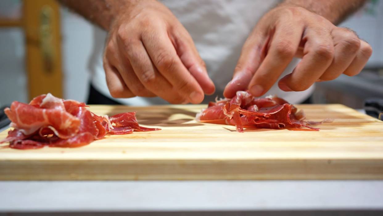 Hands arranging thin slices of cured meat on a wooden cutting board in a kitchen.